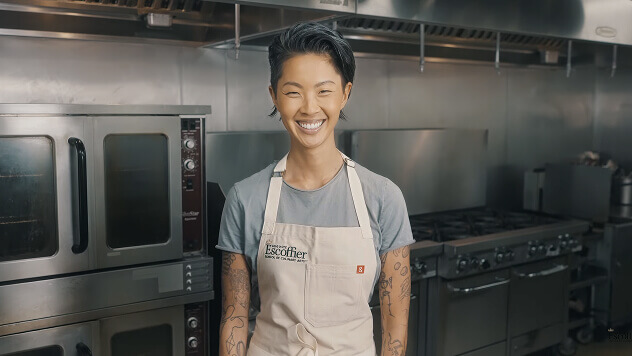 Chef Kristen Kish smiling in an Escoffier kitchen, wearing a gray shirt and Escoffier apron