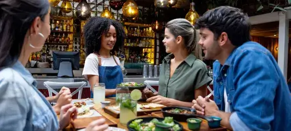 Group of friends seated at a restaurant table ordering food from a smiling waitress, with drinks and dishes on the table in a warmly lit setting