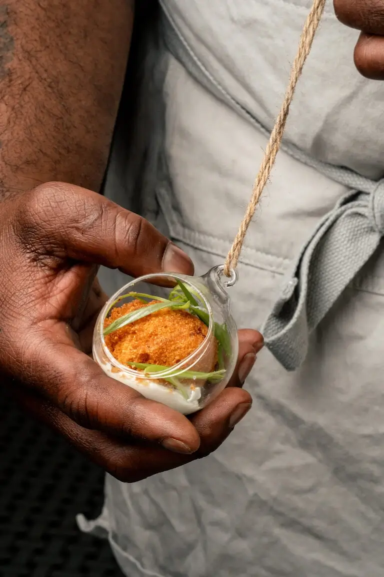 Chef Rodney holding a glass ornament with an open front filled with a hush puppy on a horseradish sauce.
