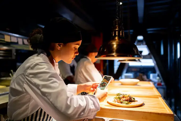 Chef in uniform taking a photo of a plated dish with her smartphone in a restaurant kitchen under warm lighting.