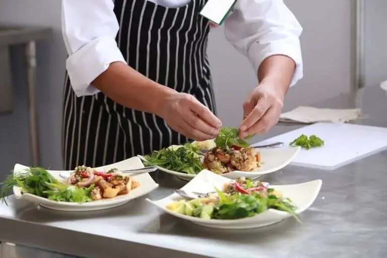 Chef plating multiple colorful dishes with fresh ingredients in a professional kitchen.