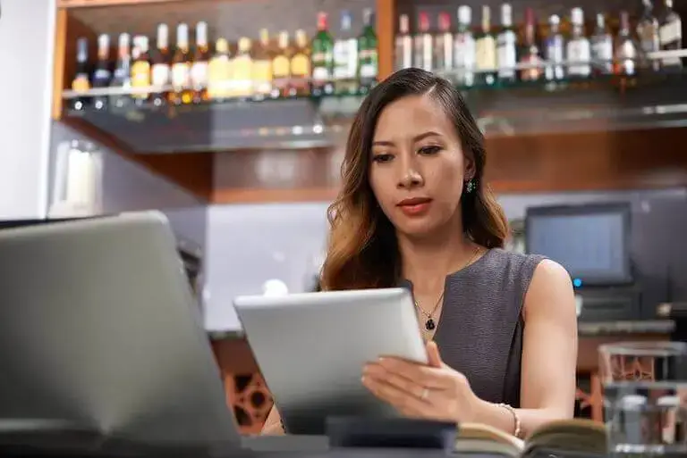 Manager dressed in a grey shirt standing in front of a bar looking at a tablet.