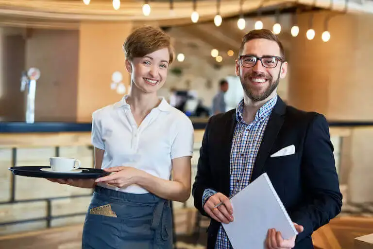 Restaurant manager and server holding a tray smile for a photo while standing in a restaurant.