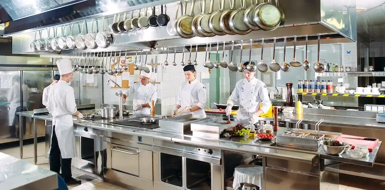Several people in chef’s uniforms work at various stations around a counter in the center of a commercial kitchen.