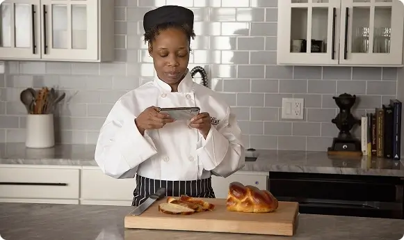 Escoffier culinary student in chef coat taking a photo of freshly baked bread in a modern kitchen