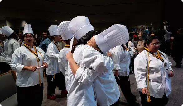 Two Escoffier culinary graduates in chef hats hug during a joyful graduation ceremony