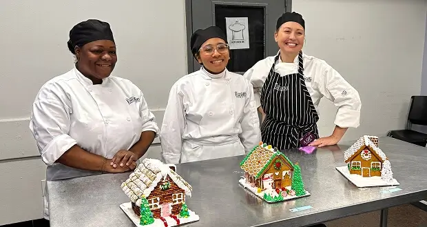 Three students in chef coats display colorful gingerbread houses during festive pastry class