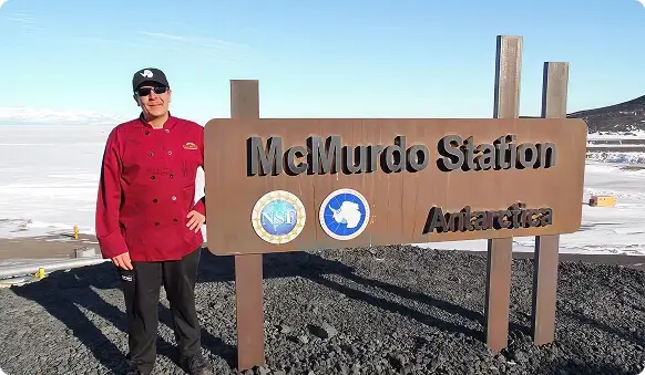Escoffier graduate Douglas Rhoades standing beside McMurdo Station sign in Antarctica with icy landscape in background