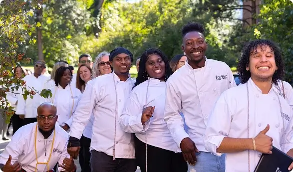 Group of Escoffier graduates in white chef coats smiling and celebrating outdoors with trees in the background.
