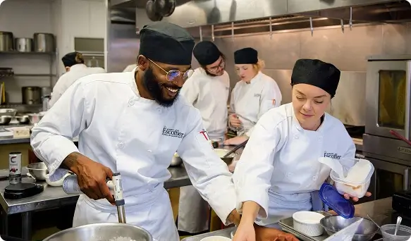 Escoffier culinary students work together in a commercial kitchen preparing dishes and sharing tools