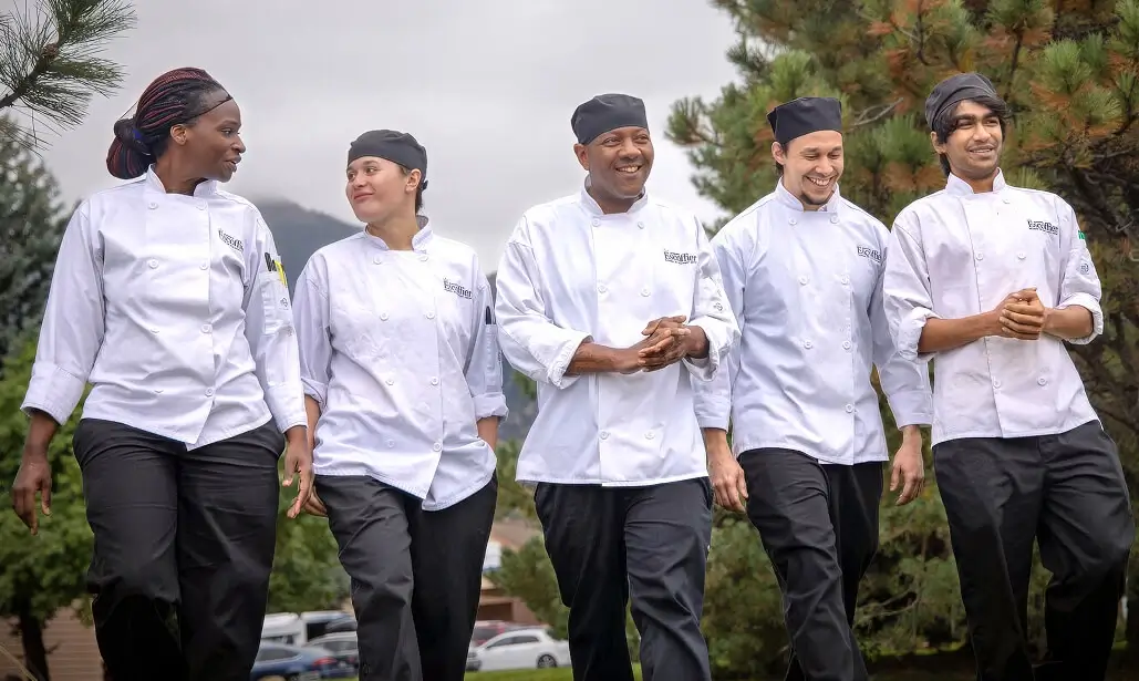 Group of five culinary students in white chef coats walking outdoors near Boulder campus, smiling and talking on a cloudy day with pine trees in the background