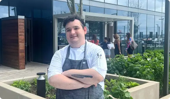 Escoffier graduate Shai Fernandez poses confidently in an urban garden setting outside a modern restaurant