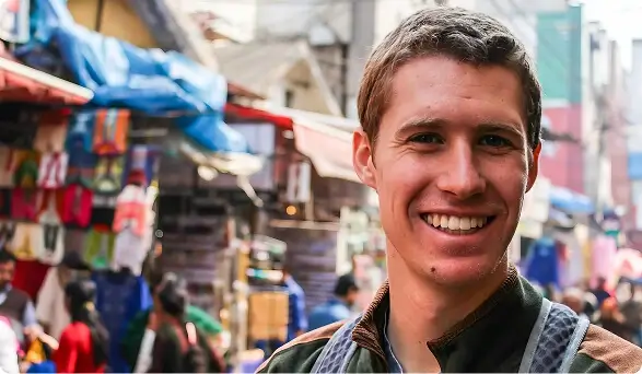 Smiling Escoffier graduate Shane Witters Hicks in street market abroad, wearing an apron and standing in front of colorful vendor stalls.