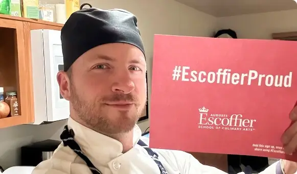 Escoffier culinary student in uniform holding a red #EscoffierProud sign in kitchen.