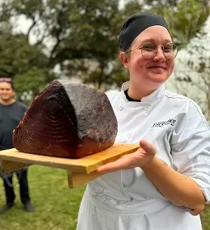 Escoffier culinary student proudly holding a large smoked meat block on a wooden tray during outdoor event