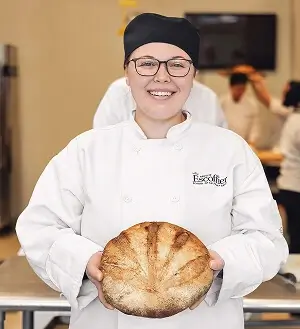 Escoffier culinary student smiling and holding a round loaf of freshly baked bread in a professional kitchen.