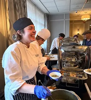 Escoffier culinary student serving food and smiling at a catered event with chef team in background