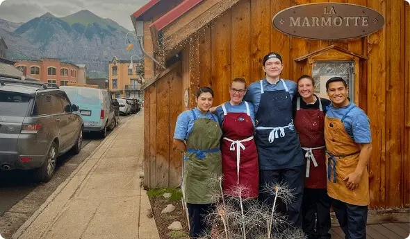 Escoffier culinary student Jackson Hussey and four other cooks in colorful aprons smile outside La Marmotte restaurant with mountain backdrop.