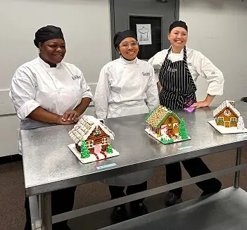 Three students in chef coats display colorful gingerbread houses during festive pastry class