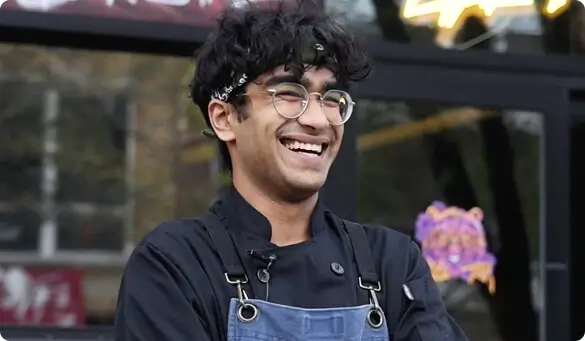 Smiling Escoffier culinary student Reshmanth Gummadi in apron and glasses standing outside a restaurant.