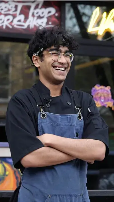 Happy Escoffier student Reshmanth Gummadi in black chef's coat and denim apron standing outside a restaurant with neon signs