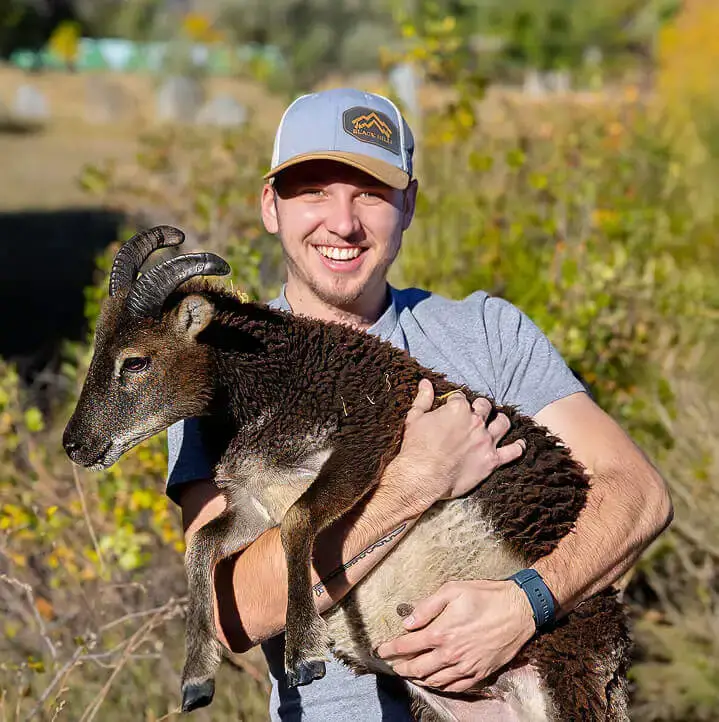 a young man in a hat smiling while holding a dark brown sheep outdoors on a sunny farm