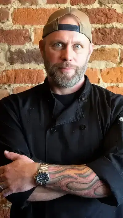 Tattooed chef Lance McWhorter wearing a black coat and backwards cap standing in front of a rustic brick wall