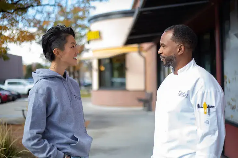 Escoffier Chef-In-Residence and Top Chef Host Kristen Kish, left, in a gray hooded sweatshirt, talks to Escoffier student Trenin Nubiru, right, who is wearing a white chef's apron.