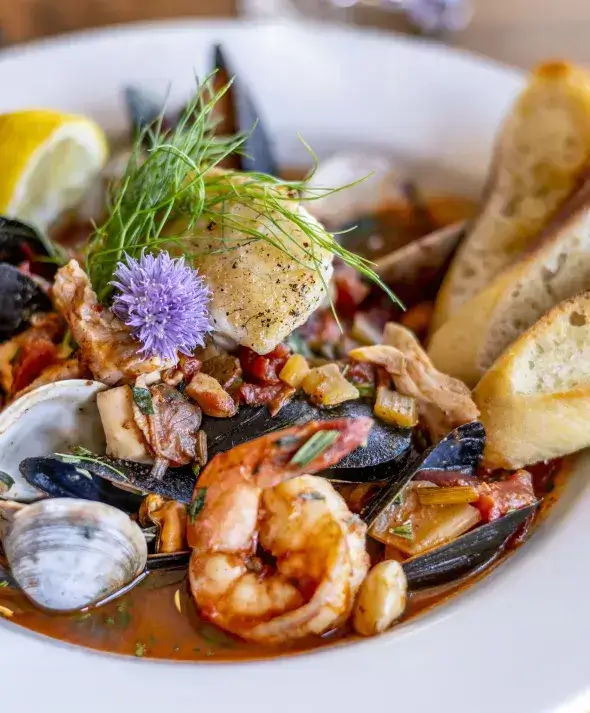 Close-up of a gourmet bouillabaisse seafood soup with shrimp, mussels, clams, white fish, fennel garnish, and toasted baguette slices served in a white bowl.