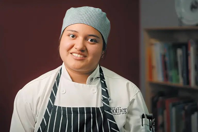 Escoffier culinary student Jacqueline in chef uniform and striped apron smiling at camera