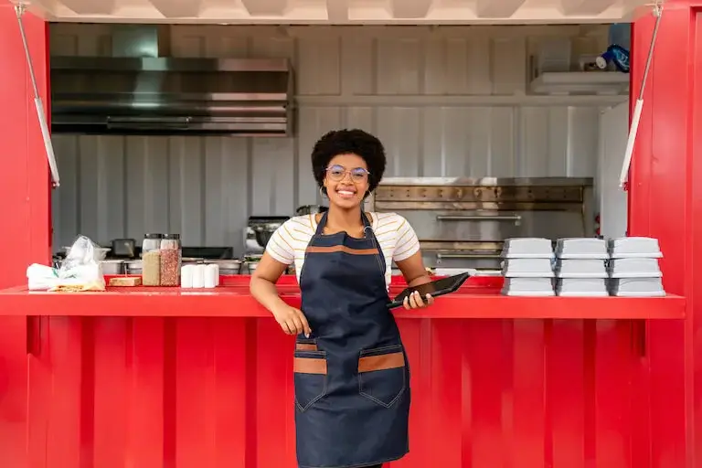 A smiling woman in an apron stands confidently in front of a red food truck counter, holding a tablet with takeaway containers stacked beside her.