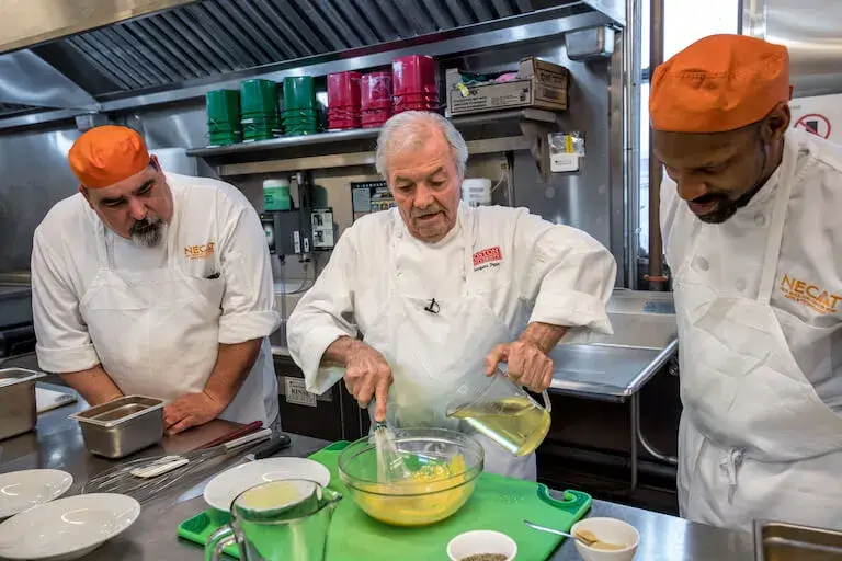 Chef Jacques Pépin pours liquid into a mixing bowl and holds a whisk while two people stand on either side of him in a teaching kitchen.