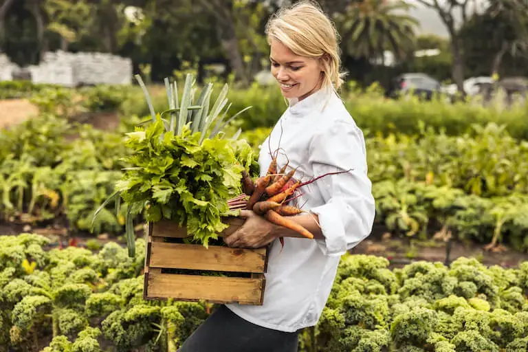 Chef in a white coat carrying a wooden crate of fresh vegetables—including celery, leeks, and carrots—while walking through a lush garden.