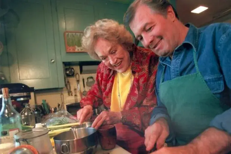 Famed chefs Julia Child and Jacques Pépin smile and are each looking down at the counter while cooking in Child's kitchen.