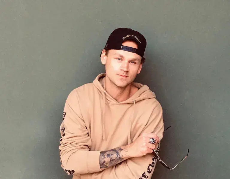 Parker Wilks-Bryant, Auguste Escoffier School of Culinary Arts graduate, wearing beige hoodie and backwards cap with arms crossed, standing against gray wall.