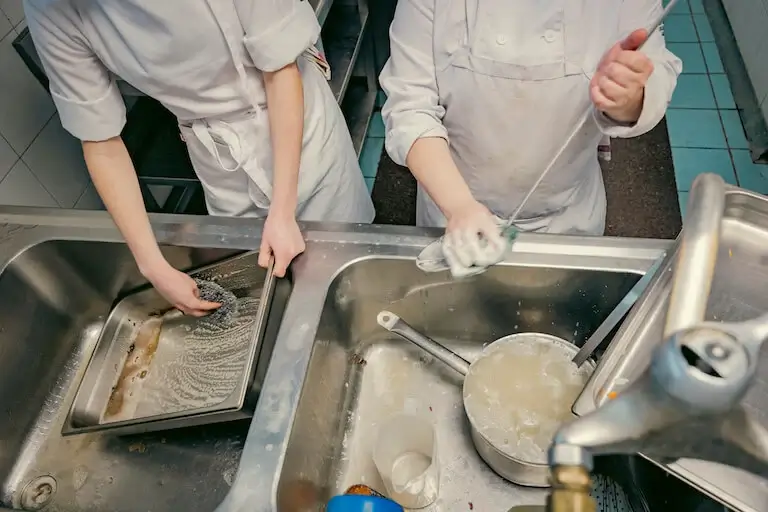 Two people in chef whites washing dishes at a commercial kitchen sink.