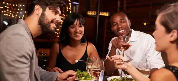 Four friends sharing laughs, wine, and salads in a warmly lit restaurant with festive lighting