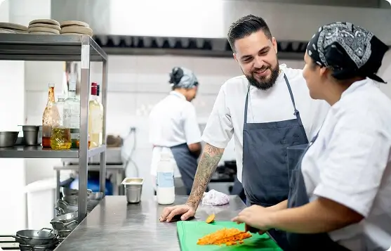 Chef mentor coaching a culinary student at a prep table in a commercial kitchen