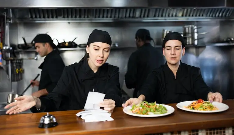 A cook places two plated dishes in the service window of a kitchen beside another cook checking a stack of service tickets.