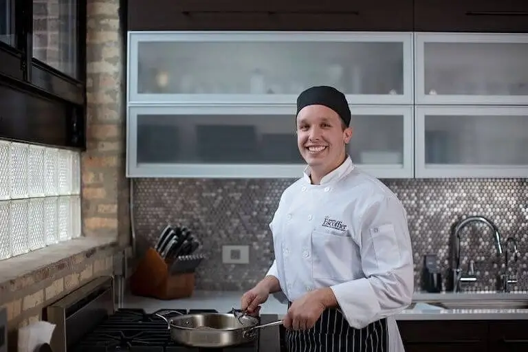 A smiling culinary student in an Escoffier chef coat stands at a stove in a home kitchen.