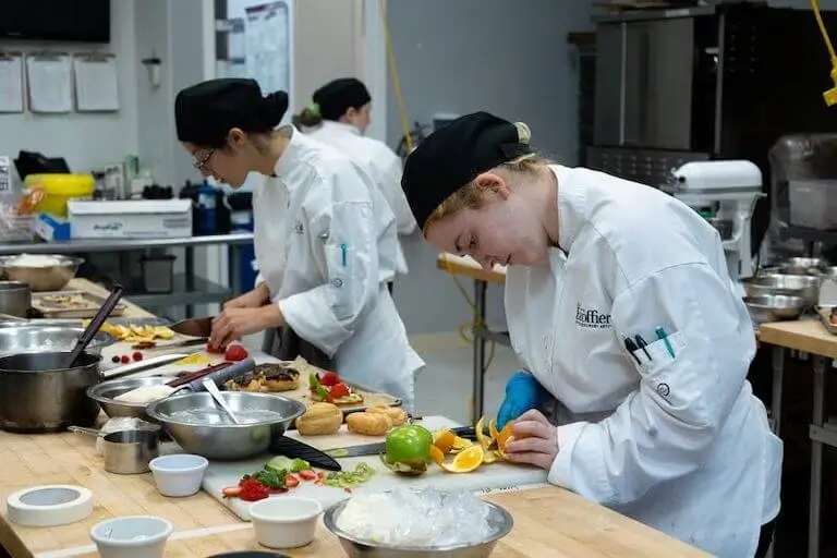 Culinary students in chef whites and hats working at individual stations in professional kitchen classroom at Escoffier campus