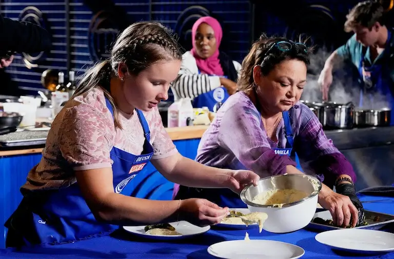 Daniela Peregrina, wearing purple shirt and blue apron, prepares a dish alongside a teammate during a MasterChef team competition.