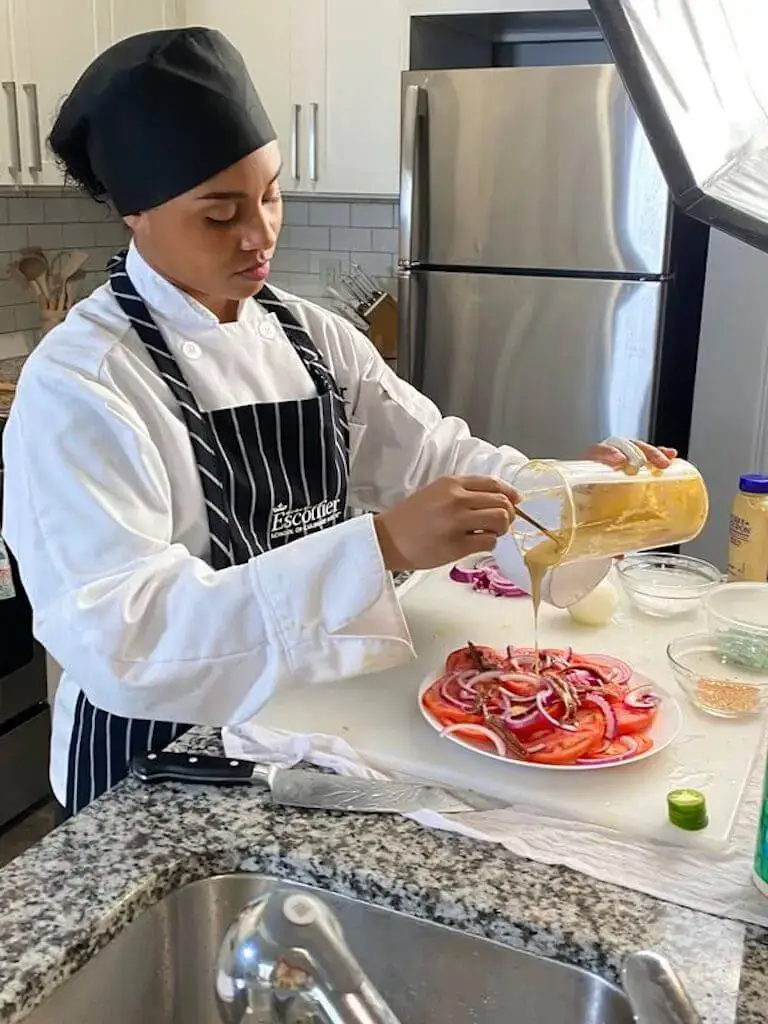 Escoffier online student in chef whites and apron preparing food in home kitchen while completing coursework