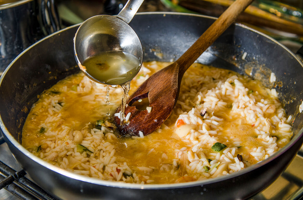 A ladle of hot broth being poured into a pan of simmering risotto, with a wooden spoon stirring the rice.