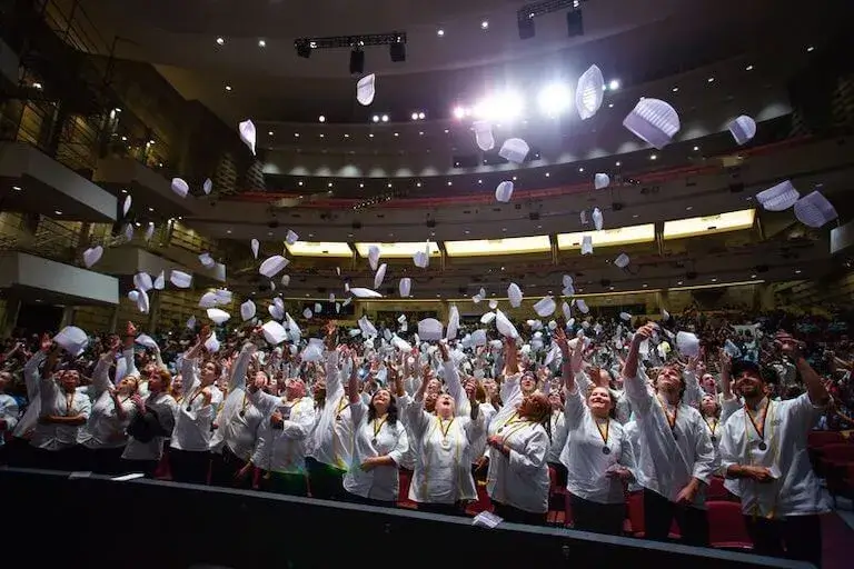 A large group of Escoffier Boulder graduates in white chef coats stand inside a theater-style auditorium, cheering and tossing their chef hats high into the air during a celebratory graduation ceremony.
