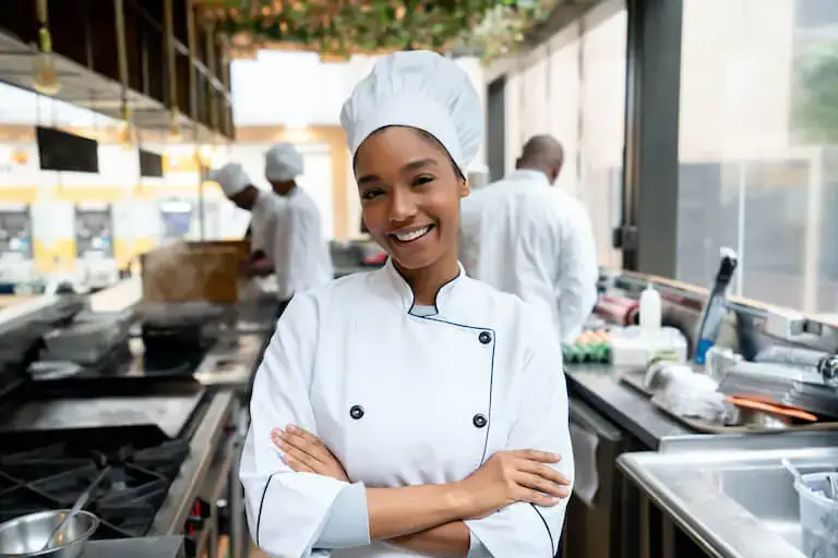 A smiling chef in a white uniform and toque stands with crossed arms in a busy commercial kitchen. The background shows other kitchen staff at stainless steel workstations.