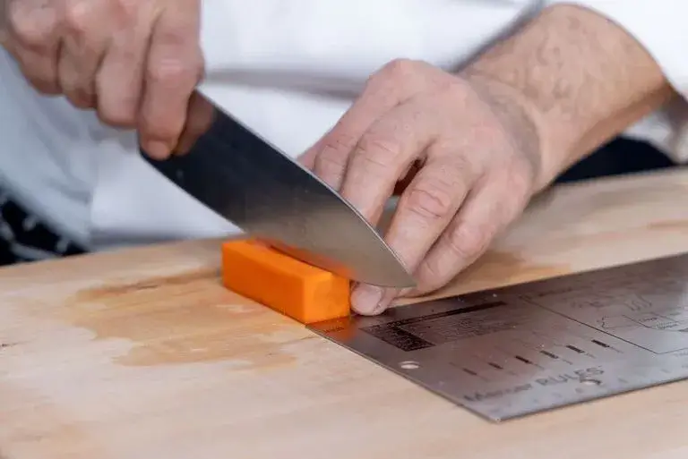 Close-up of a pair of hands making a paysenne cut on a cutting board with a a stainless steel guide nearby.