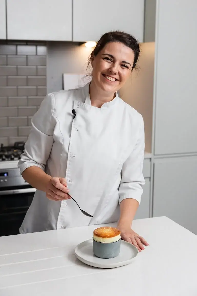Chef Marion Lancial smiles in a modern kitchen while holding a spoon beside a freshly baked dessert served in a ceramic ramekin on a white plate.