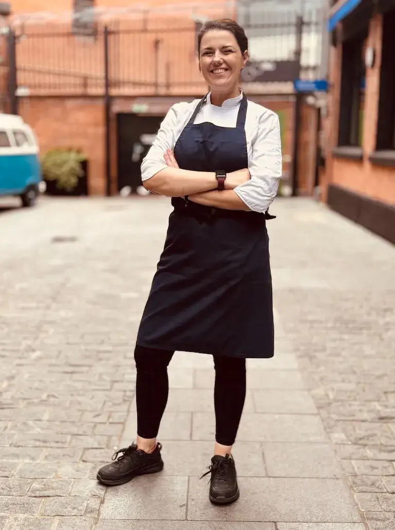 Chef Marion Lancial in chef's whites and black apron standing confidently with arms crossed on cobblestone street in Belfast, Northern Ireland