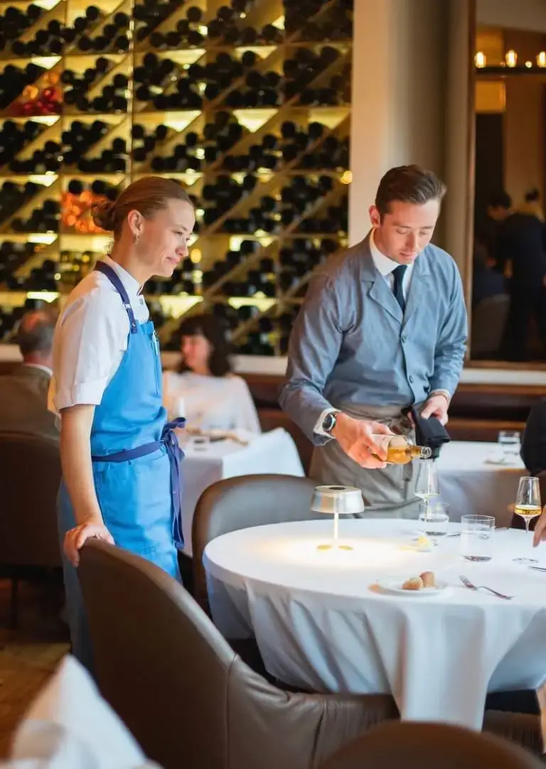 Sommelier Connor Fowler pours wine at a table in an upscale restaurant dining room with an illuminated wine wall in the background